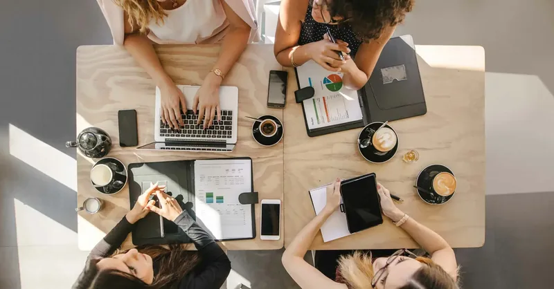 Vue de dessus de quatre personnes en réunion autour d'une table, avec ordinateurs, tablettes et cafés